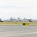 Two military jet aircraft, appearing to be F/A-18 Hornets, are operating on a runway at a military airbase. A yellow taxiway sign reading 'F1' with directional arrows is prominently visible in the foreground. The background shows an air traffic control tower, base infrastructure, and mountainous terrain.