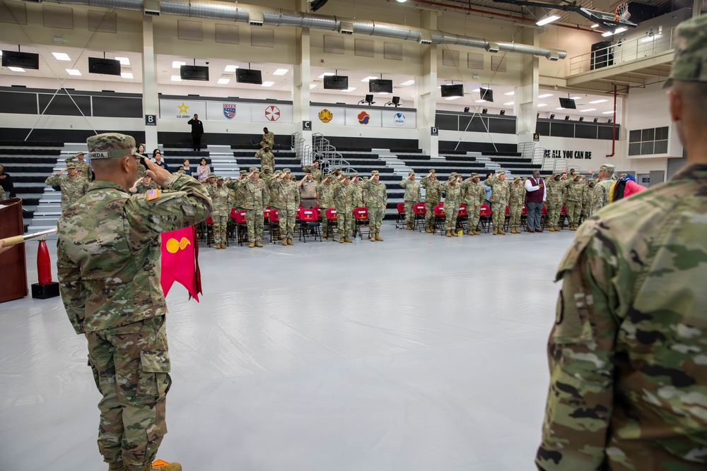 A U.S. Army military ceremony is taking place inside a large indoor gymnasium or sports facility. Soldiers in Army Combat Uniforms (ACU/OCP pattern) are standing in formation and rendering salutes, with a guidon bearer holding a red unit guidon. The venue displays multiple military unit insignia banners and the U.S. Army slogan 'BE ALL YOU CAN BE' on the wall.