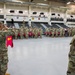 A U.S. Army military ceremony is taking place inside a large indoor gymnasium or sports facility. Soldiers in Army Combat Uniforms (ACU/OCP pattern) are standing in formation and rendering salutes, with a guidon bearer holding a red unit guidon. The venue displays multiple military unit insignia banners and the U.S. Army slogan 'BE ALL YOU CAN BE' on the wall.