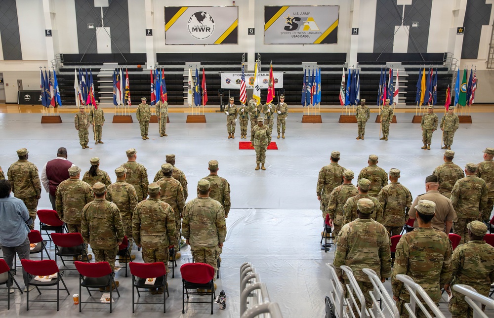 A U.S. Army military ceremony is taking place inside a gymnasium at USAG Daegu, South Korea, identified by signage for U.S. Army MWR and Sports-Fitness-Aquatics. The event appears to be a change of command or unit ceremony for the 6th Ordnance Battalion, with soldiers in formation facing a central color guard holding multiple flags. The venue features bleachers, state and unit flags displayed on stands, and an audience of soldiers and civilians seated and standing.