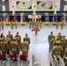 A U.S. Army military ceremony is taking place inside a gymnasium at USAG Daegu, South Korea, identified by signage for U.S. Army MWR and Sports-Fitness-Aquatics. The event appears to be a change of command or unit ceremony for the 6th Ordnance Battalion, with soldiers in formation facing a central color guard holding multiple flags. The venue features bleachers, state and unit flags displayed on stands, and an audience of soldiers and civilians seated and standing.