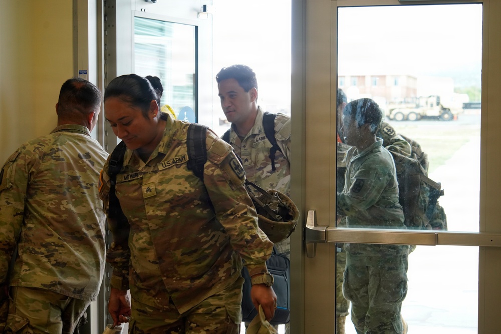 A group of U.S. Army soldiers in OCP (Operational Camouflage Pattern) uniforms are entering or exiting a building through glass doors. The soldiers are carrying backpacks and gear, suggesting a movement or transit operation. A military vehicle and outdoor installation area are visible through the glass doors in the background.