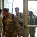 A group of U.S. Army soldiers in OCP (Operational Camouflage Pattern) uniforms are entering or exiting a building through glass doors. The soldiers are carrying backpacks and gear, suggesting a movement or transit operation. A military vehicle and outdoor installation area are visible through the glass doors in the background.