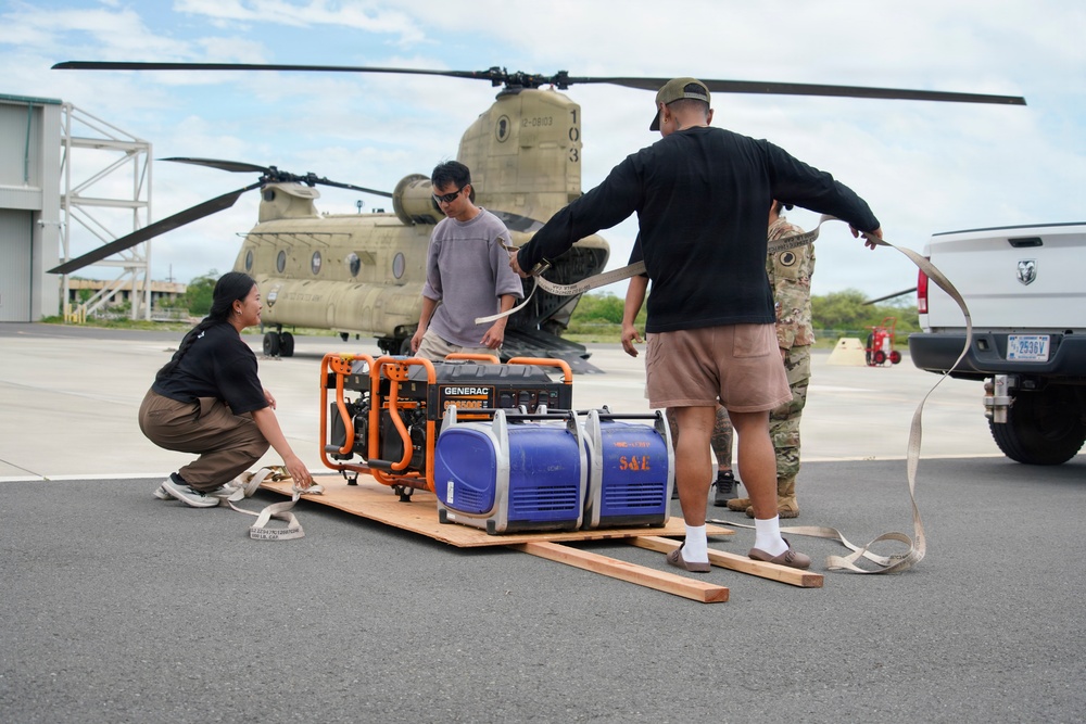 A group of civilians and at least one military personnel are preparing generators for transport via a CH-47 Chinook helicopter on a tarmac. The individuals are securing two portable generators — a Generac and a blue unit marked 'S&amp;E' — on a wooden pallet using cargo straps. This appears to be a disaster relief or emergency supply operation.