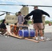A group of civilians and at least one military personnel are preparing generators for transport via a CH-47 Chinook helicopter on a tarmac. The individuals are securing two portable generators — a Generac and a blue unit marked 'S&amp;E' — on a wooden pallet using cargo straps. This appears to be a disaster relief or emergency supply operation.