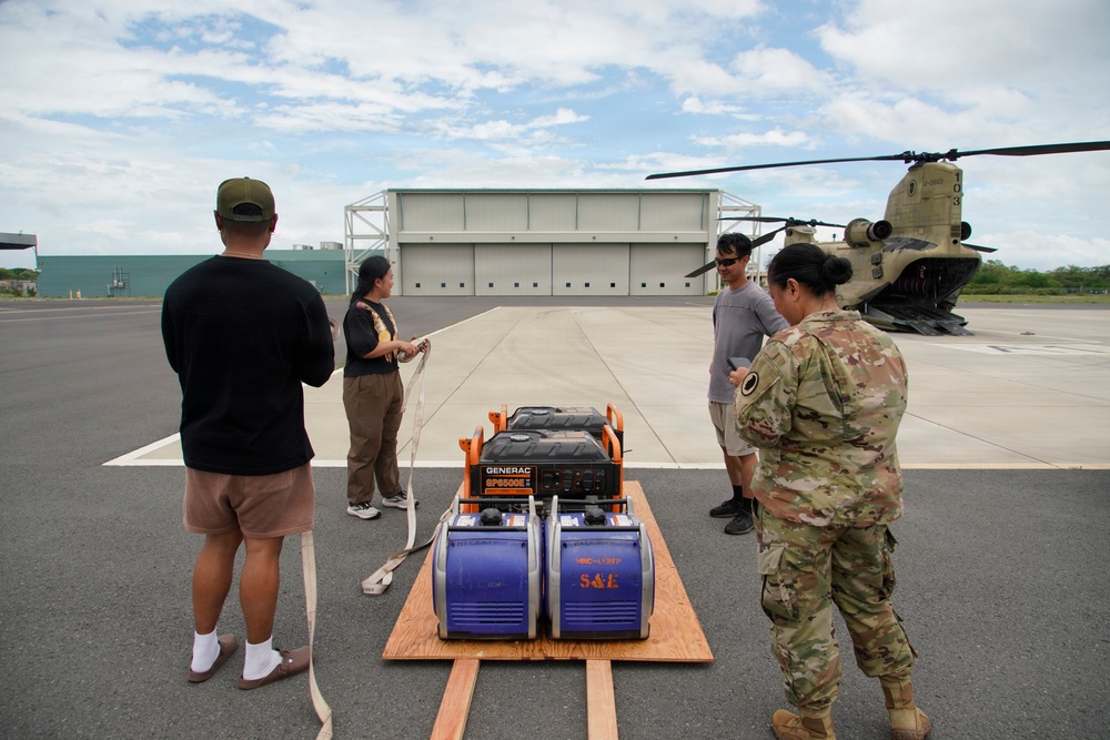 A military logistics operation on an airfield tarmac where personnel are preparing generators for transport. A CH-47 Chinook helicopter is parked in the background near a hangar, suggesting the generators will be airlifted. The scene appears to be a disaster relief or emergency supply operation, with both military and civilian personnel present.