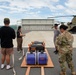 A military logistics operation on an airfield tarmac where personnel are preparing generators for transport. A CH-47 Chinook helicopter is parked in the background near a hangar, suggesting the generators will be airlifted. The scene appears to be a disaster relief or emergency supply operation, with both military and civilian personnel present.