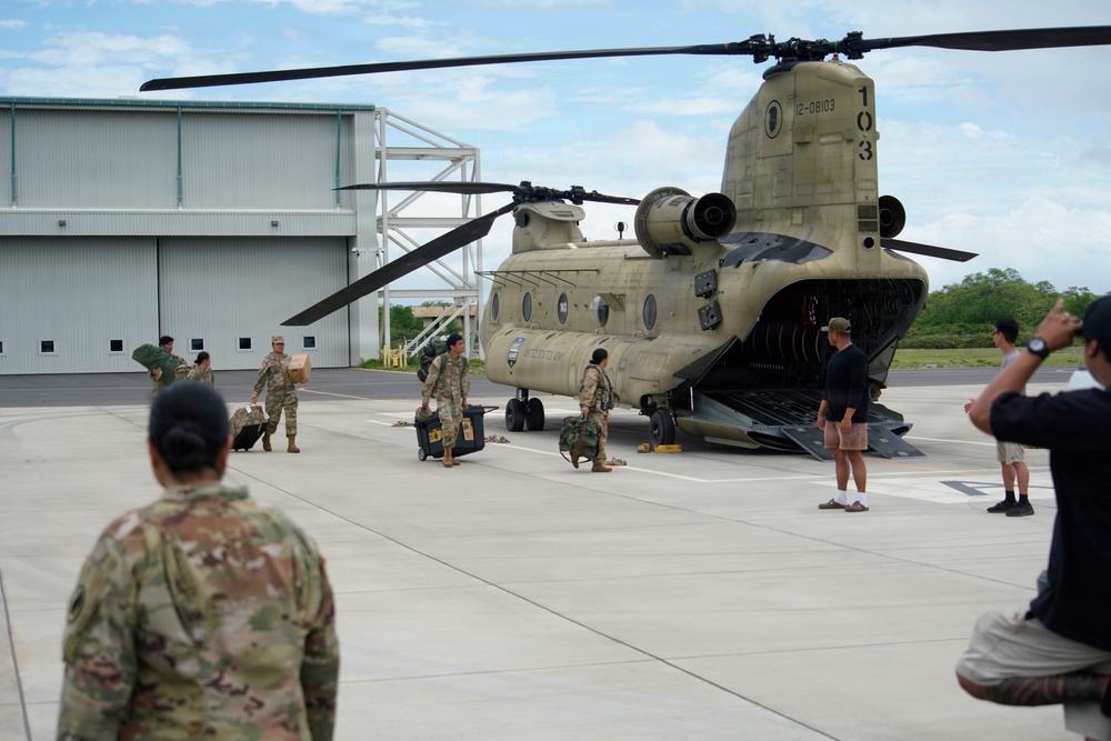 A U.S. Army CH-47 Chinook helicopter (tail number 12-08103, marked '103') is parked on a tarmac with its rear ramp open. Military personnel in camouflage uniforms are unloading or loading cargo and equipment from the aircraft. Several civilians are also present on the tarmac, with one appearing to photograph the scene.