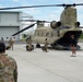A U.S. Army CH-47 Chinook helicopter (tail number 12-08103, marked '103') is parked on a tarmac with its rear ramp open. Military personnel in camouflage uniforms are unloading or loading cargo and equipment from the aircraft. Several civilians are also present on the tarmac, with one appearing to photograph the scene.