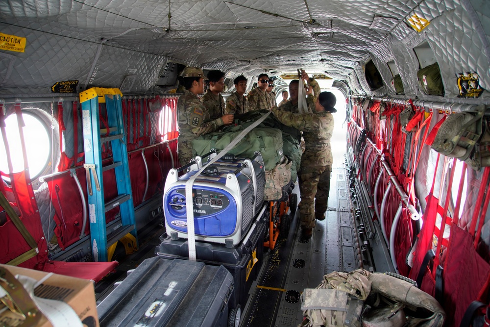 Military personnel in camouflage uniforms are loading and securing cargo inside what appears to be a CH-47 Chinook helicopter. The interior shows multiple soldiers working together to strap down equipment including a Yamaha generator and other gear. The aircraft interior features characteristic red cargo netting, quilted insulation panels, and circular windows.