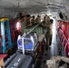 Military personnel in camouflage uniforms are loading and securing cargo inside what appears to be a CH-47 Chinook helicopter. The interior shows multiple soldiers working together to strap down equipment including a Yamaha generator and other gear. The aircraft interior features characteristic red cargo netting, quilted insulation panels, and circular windows.