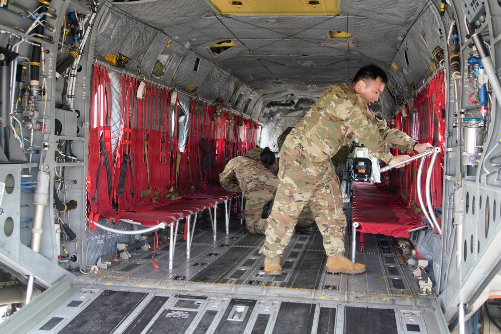 Two military personnel in camouflage uniforms are working inside the cargo hold of a large military helicopter, likely a CH-47 Chinook. They appear to be setting up or configuring red canvas litter/stretcher systems along the interior walls, suggesting preparation for a medevac or aeromedical evacuation mission. The interior shows the characteristic gray metal structure with visible hydraulic lines, cables, and equipment typical of a heavy-lift military transport helicopter.
