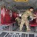 Two military personnel in camouflage uniforms are working inside the cargo hold of a large military helicopter, likely a CH-47 Chinook. They appear to be setting up or configuring red canvas litter/stretcher systems along the interior walls, suggesting preparation for a medevac or aeromedical evacuation mission. The interior shows the characteristic gray metal structure with visible hydraulic lines, cables, and equipment typical of a heavy-lift military transport helicopter.