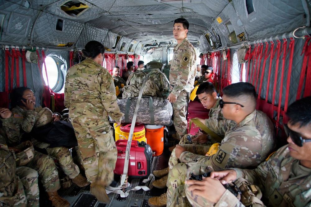 Military personnel in OCP (Operational Camouflage Pattern) uniforms are seated and standing inside what appears to be a CH-47 Chinook helicopter. The soldiers are transporting equipment including a red Honda generator, a blue water cooler, and military duffel bags secured with cargo straps. The scene suggests a logistics or disaster relief operation given the civilian utility equipment present.