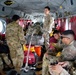 Military personnel in OCP (Operational Camouflage Pattern) uniforms are seated and standing inside what appears to be a CH-47 Chinook helicopter. The soldiers are transporting equipment including a red Honda generator, a blue water cooler, and military duffel bags secured with cargo straps. The scene suggests a logistics or disaster relief operation given the civilian utility equipment present.