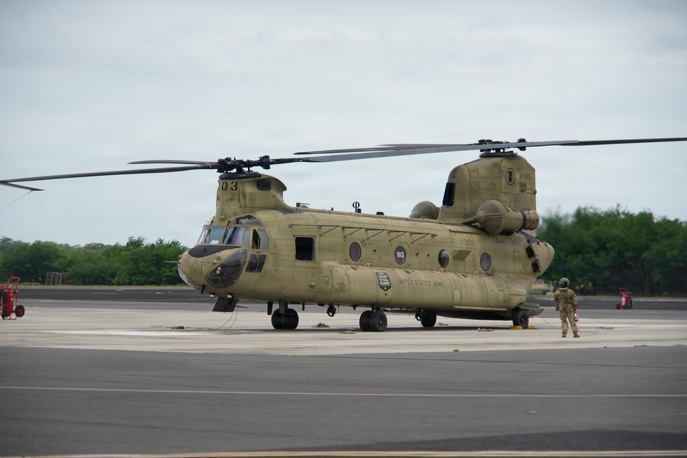 A United States Army CH-47 Chinook tandem-rotor helicopter is parked on a tarmac or airfield apron. The aircraft bears 'UNITED STATES ARMY' and 'ARMY GUARD' markings and is painted in standard olive drab. A single personnel member in flight gear stands near the rear of the aircraft, and fire suppression equipment is visible on both sides of the frame.