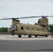 A United States Army CH-47 Chinook tandem-rotor helicopter is parked on a tarmac or airfield apron. The aircraft bears 'UNITED STATES ARMY' and 'ARMY GUARD' markings and is painted in standard olive drab. A single personnel member in flight gear stands near the rear of the aircraft, and fire suppression equipment is visible on both sides of the frame.