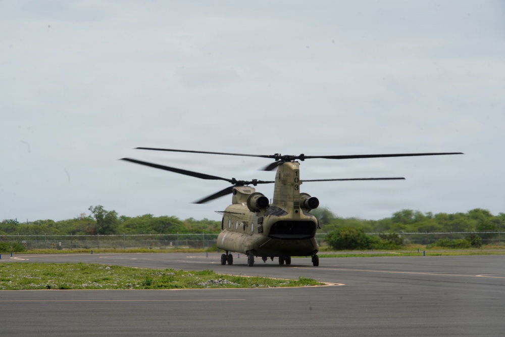 A military CH-47 Chinook tandem-rotor helicopter is on a paved tarmac or helipad with its rotors spinning, suggesting it is either preparing for takeoff or has just landed. The helicopter is painted in olive drab/army green camouflage livery. The setting appears to be a small airfield or military installation surrounded by tropical or subtropical vegetation.