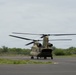 A military CH-47 Chinook tandem-rotor helicopter is on a paved tarmac or helipad with its rotors spinning, suggesting it is either preparing for takeoff or has just landed. The helicopter is painted in olive drab/army green camouflage livery. The setting appears to be a small airfield or military installation surrounded by tropical or subtropical vegetation.