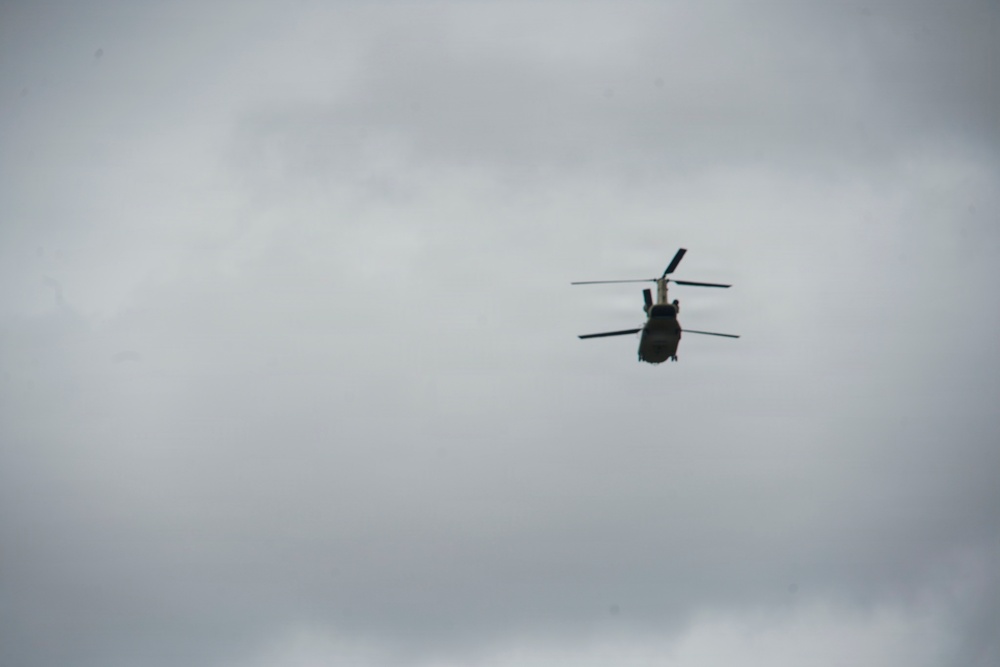 A tandem-rotor helicopter, consistent with a CH-47 Chinook design, is photographed in flight against an overcast, grey sky. The aircraft appears dark/olive in color and is viewed from a slightly below-rear angle. The image was taken at a distance, resulting in limited fine detail of the aircraft.