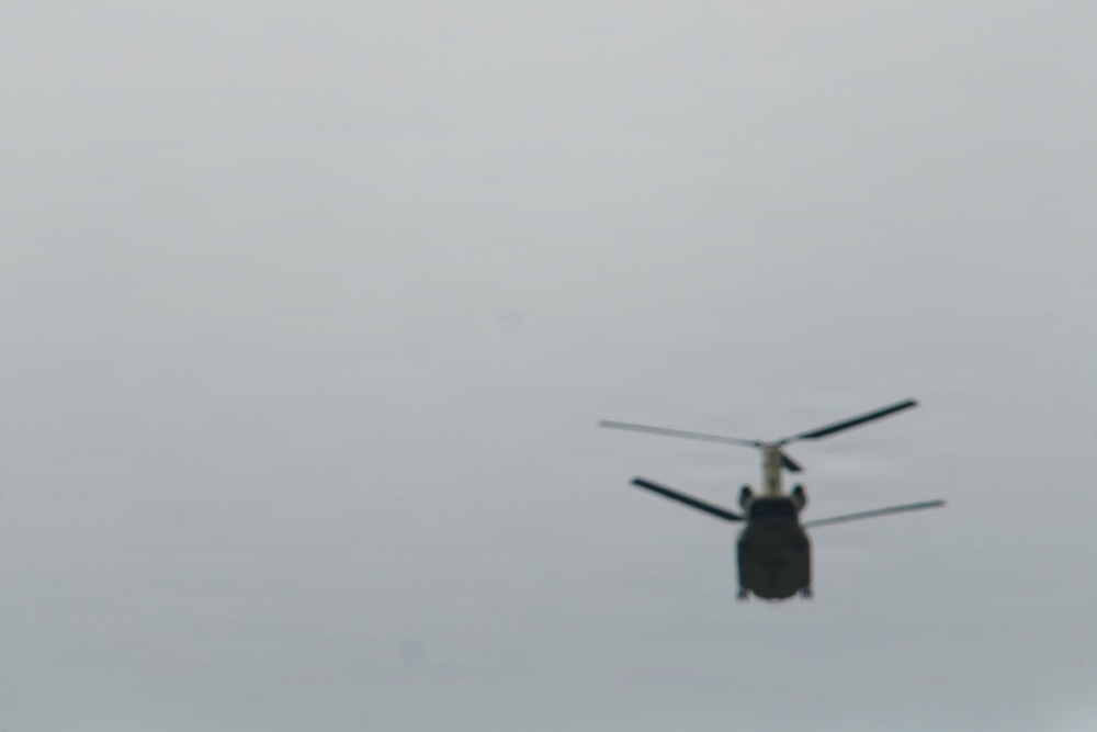 A blurry photograph captures a tandem-rotor helicopter in flight against an overcast gray sky. The aircraft appears to be a heavy-lift helicopter, likely a Boeing CH-47 Chinook, identifiable by its distinctive dual-rotor configuration. The image is taken from below and at a distance, resulting in significant blur and limited detail.