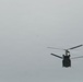 A blurry photograph captures a tandem-rotor helicopter in flight against an overcast gray sky. The aircraft appears to be a heavy-lift helicopter, likely a Boeing CH-47 Chinook, identifiable by its distinctive dual-rotor configuration. The image is taken from below and at a distance, resulting in significant blur and limited detail.
