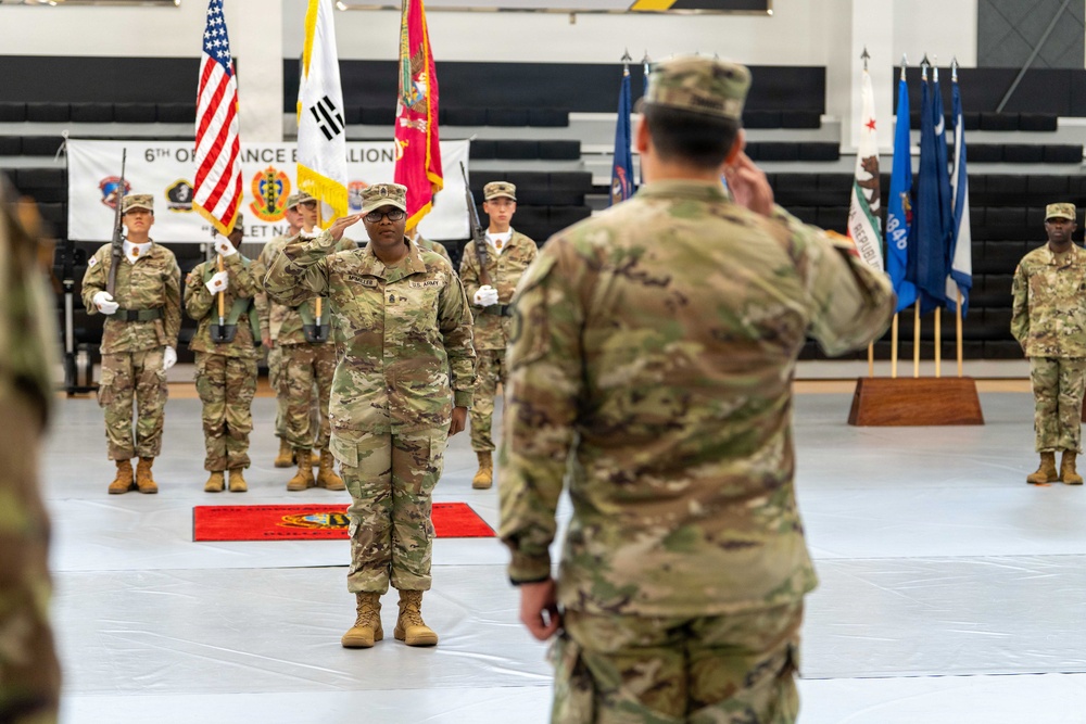 A formal U.S. Army military ceremony is taking place inside an indoor gymnasium or arena. Two soldiers are exchanging salutes in the foreground, while a color guard stands at attention in the background holding multiple flags including the U.S. flag and South Korean flag. A banner in the background identifies this as a 6th Ordnance Battalion event.