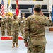 A formal U.S. Army military ceremony is taking place inside an indoor gymnasium or arena. Two soldiers are exchanging salutes in the foreground, while a color guard stands at attention in the background holding multiple flags including the U.S. flag and South Korean flag. A banner in the background identifies this as a 6th Ordnance Battalion event.