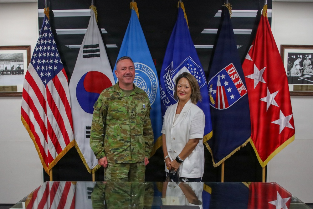 Two individuals pose for a formal photograph in what appears to be a military command facility. One person is wearing a military camouflage uniform and the other is dressed in civilian attire (white blazer). They are standing in front of an array of official flags representing multiple military commands.