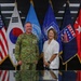 Two individuals pose for a formal photograph in what appears to be a military command facility. One person is wearing a military camouflage uniform and the other is dressed in civilian attire (white blazer). They are standing in front of an array of official flags representing multiple military commands.