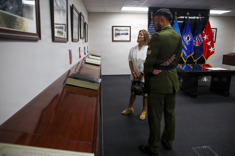 A military officer in olive green dress uniform with decorative sash is standing alongside a civilian woman in a white blazer, both viewing framed photographs and documents displayed on a wooden ledge along a wall. The setting appears to be an official military office or ceremonial room, evidenced by multiple organizational flags visible in the background. The flags include what appear to be U.S. Army command flags, a South Korean flag element, and a U.S. national flag.