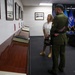 A military officer in olive green dress uniform with decorative sash is standing alongside a civilian woman in a white blazer, both viewing framed photographs and documents displayed on a wooden ledge along a wall. The setting appears to be an official military office or ceremonial room, evidenced by multiple organizational flags visible in the background. The flags include what appear to be U.S. Army command flags, a South Korean flag element, and a U.S. national flag.