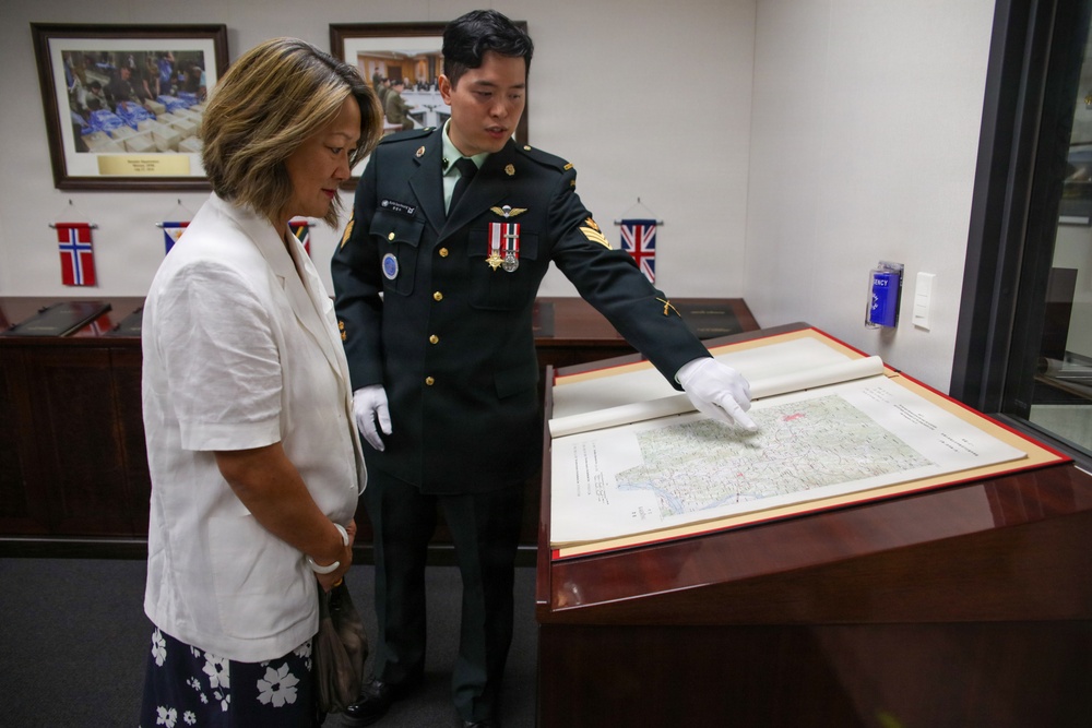 A uniformed military officer wearing white conservation gloves is presenting a large historical map to a civilian woman in a formal room. The officer is pointing to specific locations on the map, which is displayed in a red-bordered presentation case on a wooden table. The setting appears to be an official military or archival facility, with framed photographs and international flags visible in the background.