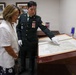 A uniformed military officer wearing white conservation gloves is presenting a large historical map to a civilian woman in a formal room. The officer is pointing to specific locations on the map, which is displayed in a red-bordered presentation case on a wooden table. The setting appears to be an official military or archival facility, with framed photographs and international flags visible in the background.