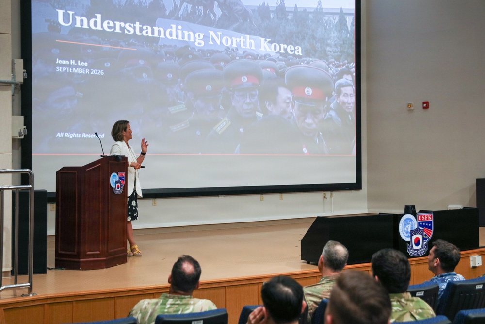 A woman is delivering a presentation titled 'Understanding North Korea' by Jean H. Lee, dated September 2026, at what appears to be a U.S. Forces Korea (USFK) military installation. She stands at a wooden podium on a stage while an audience that includes military personnel in camouflage uniforms listens. The USFK emblem is visible on both the podium and a display stand on the stage.