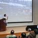 A woman is delivering a presentation titled 'Understanding North Korea' by Jean H. Lee, dated September 2026, at what appears to be a U.S. Forces Korea (USFK) military installation. She stands at a wooden podium on a stage while an audience that includes military personnel in camouflage uniforms listens. The USFK emblem is visible on both the podium and a display stand on the stage.