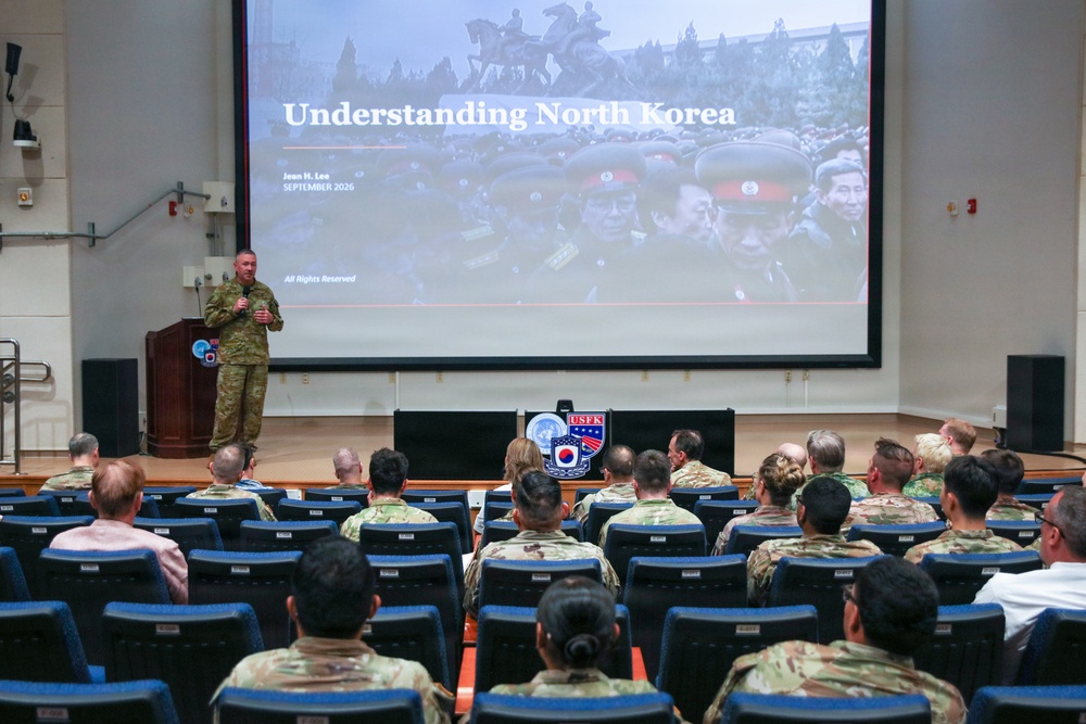 A military briefing is taking place in an auditorium-style venue where a uniformed speaker addresses an audience of military personnel. The presentation slide visible on the large screen is titled 'Understanding North Korea' by Jean H. Lee, dated September 2026. A USFK (United States Forces Korea) emblem is prominently displayed on a podium in the center of the stage.