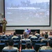 A military briefing is taking place in an auditorium-style venue where a uniformed speaker addresses an audience of military personnel. The presentation slide visible on the large screen is titled 'Understanding North Korea' by Jean H. Lee, dated September 2026. A USFK (United States Forces Korea) emblem is prominently displayed on a podium in the center of the stage.