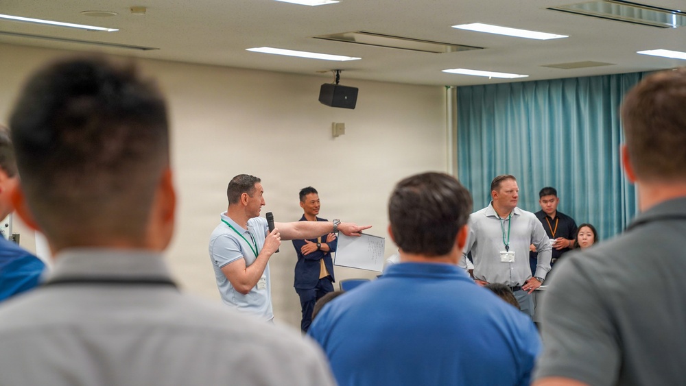 A group meeting or briefing is taking place in a conference or training room. A man holding a microphone appears to be speaking or presenting while gesturing toward a whiteboard or document, with several other individuals standing nearby. The audience is viewed from behind, suggesting a formal or semi-formal group session.