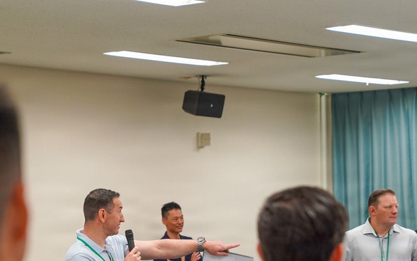 A group meeting or briefing is taking place in a conference or training room. A man holding a microphone appears to be speaking or presenting while gesturing toward a whiteboard or document, with several other individuals standing nearby. The audience is viewed from behind, suggesting a formal or semi-formal group session.
