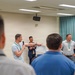 A group meeting or briefing is taking place in a conference or training room. A man holding a microphone appears to be speaking or presenting while gesturing toward a whiteboard or document, with several other individuals standing nearby. The audience is viewed from behind, suggesting a formal or semi-formal group session.