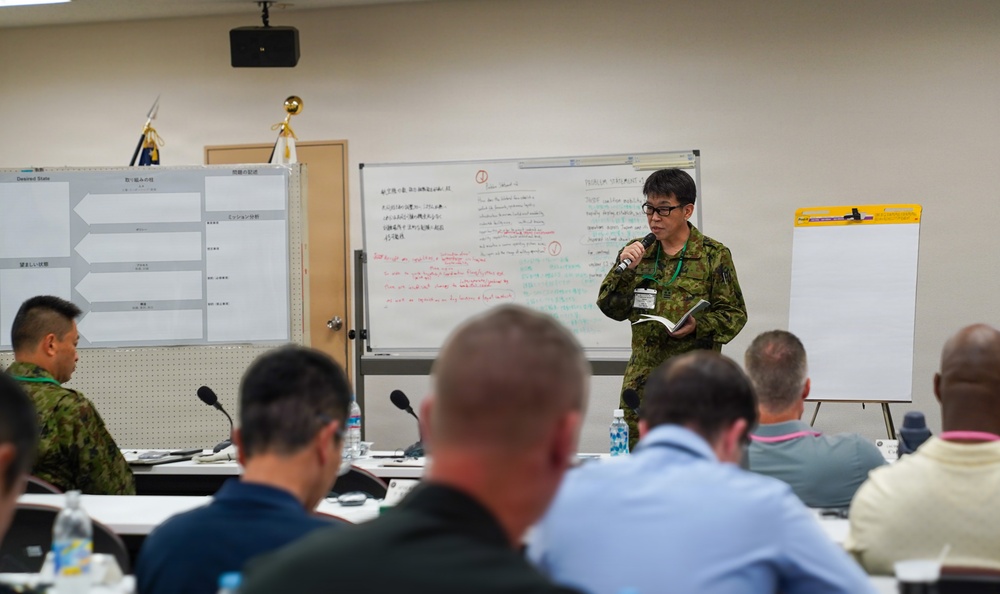 A military briefing or training session is taking place in a conference room, with a uniformed presenter holding a microphone and notebook addressing a seated audience. The setting appears to be a joint military exercise or workshop, evidenced by the mix of uniforms and civilian attire among attendees. Whiteboards behind the presenter contain both Japanese and English text, suggesting a bilateral or multinational military collaboration.