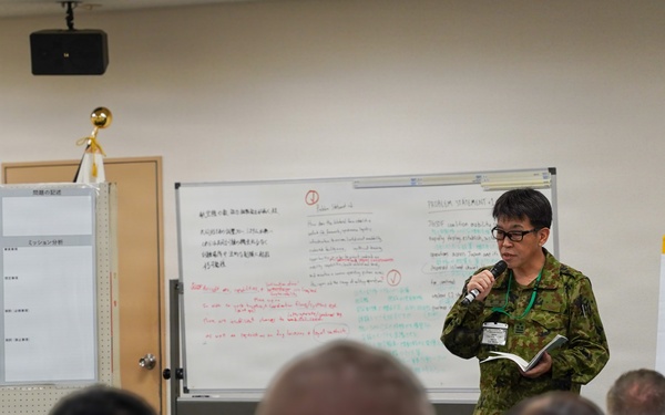 A military briefing or training session is taking place in a conference room, with a uniformed presenter holding a microphone and notebook addressing a seated audience. The setting appears to be a joint military exercise or workshop, evidenced by the mix of uniforms and civilian attire among attendees. Whiteboards behind the presenter contain both Japanese and English text, suggesting a bilateral or multinational military collaboration.