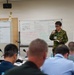 A military briefing or training session is taking place in a conference room, with a uniformed presenter holding a microphone and notebook addressing a seated audience. The setting appears to be a joint military exercise or workshop, evidenced by the mix of uniforms and civilian attire among attendees. Whiteboards behind the presenter contain both Japanese and English text, suggesting a bilateral or multinational military collaboration.