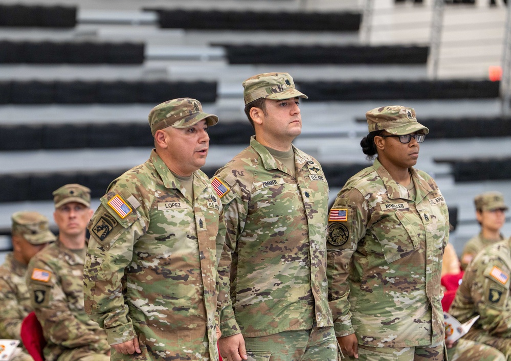 Three U.S. Army soldiers stand at attention in the foreground during what appears to be a formal military ceremony or formation. Their name tapes read 'Lopez,' 'Zimmer,' and 'McMiller,' and all wear the Operational Camouflage Pattern (OCP) uniform with U.S. flag patches. Additional soldiers are visible seated in the background near bleachers.