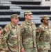 Three U.S. Army soldiers stand at attention in the foreground during what appears to be a formal military ceremony or formation. Their name tapes read 'Lopez,' 'Zimmer,' and 'McMiller,' and all wear the Operational Camouflage Pattern (OCP) uniform with U.S. flag patches. Additional soldiers are visible seated in the background near bleachers.