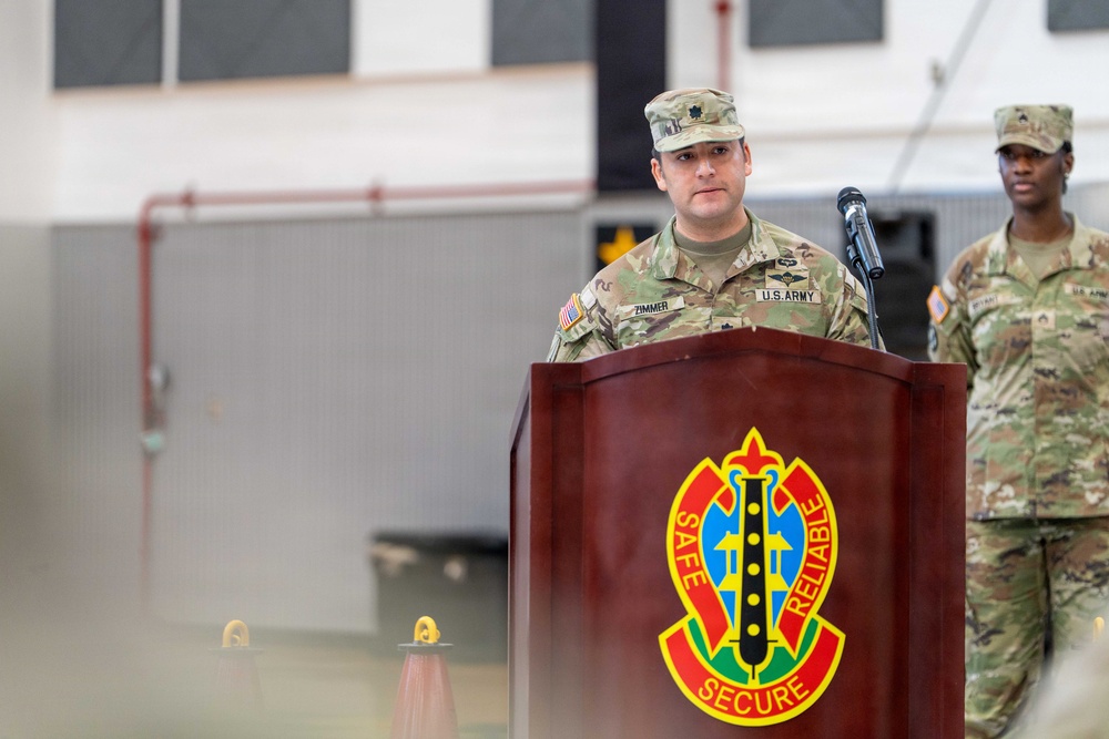 A U.S. Army soldier identified by name tape as 'ZIMMER' stands at a wooden podium bearing a military unit insignia with the motto 'Safe, Secure, Reliable.' The soldier appears to be delivering a speech or remarks at an official military ceremony or event. A second soldier stands in the background to the right.