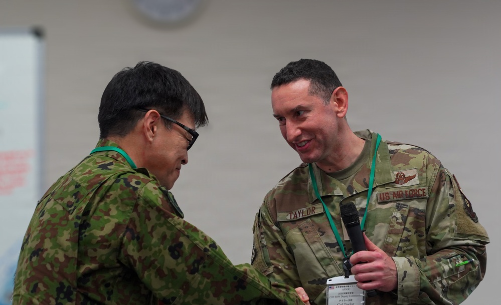 Two military personnel in camouflage uniforms are engaged in a handshake at what appears to be a formal military event or conference. One individual is wearing a US Air Force uniform with the name tape 'TAYLOR' and holding a microphone, while the other appears to be wearing a Japanese Self-Defense Force uniform. Both are wearing green lanyards/badges consistent with a conference or joint exercise setting.