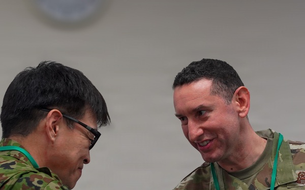 Two military personnel in camouflage uniforms are engaged in a handshake at what appears to be a formal military event or conference. One individual is wearing a US Air Force uniform with the name tape 'TAYLOR' and holding a microphone, while the other appears to be wearing a Japanese Self-Defense Force uniform. Both are wearing green lanyards/badges consistent with a conference or joint exercise setting.