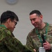 Two military personnel in camouflage uniforms are engaged in a handshake at what appears to be a formal military event or conference. One individual is wearing a US Air Force uniform with the name tape 'TAYLOR' and holding a microphone, while the other appears to be wearing a Japanese Self-Defense Force uniform. Both are wearing green lanyards/badges consistent with a conference or joint exercise setting.